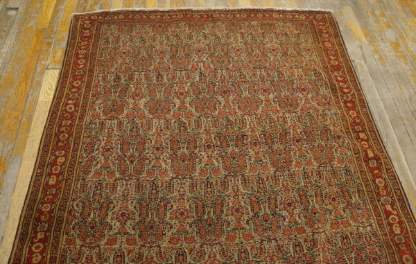 Traditional red and tan rug with dense boteh design and floral border rests on worn wooden flooring.