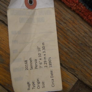 A close-up shows a wired rug tag and high-pile red and dark blue rug edge on wood flooring.