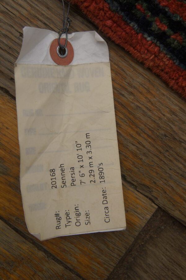 A close-up shows a wired rug tag and high-pile red and dark blue rug edge on wood flooring.