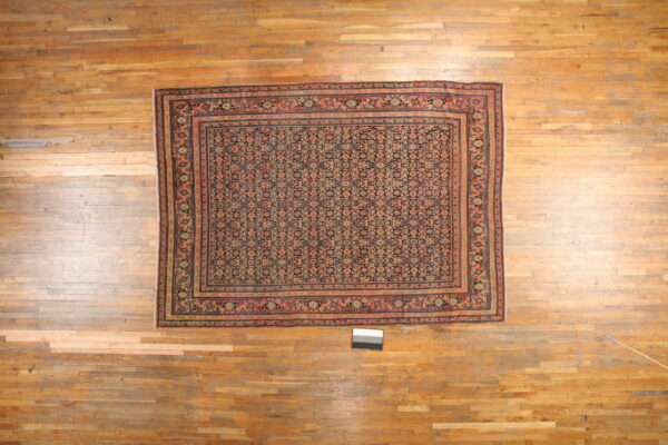 Densely patterned rug featuring coral, teal, and dark blue motifs displayed on light, worn wood flooring.