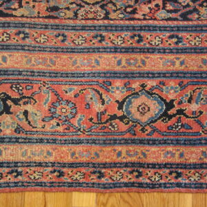 Red, navy, and beige rug border shows stylized floral and geometric bands resting on wood floor.