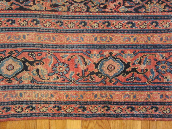 Red, navy, and beige rug border shows stylized floral and geometric bands resting on wood floor.