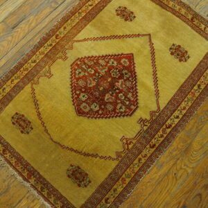 Low-pile yellow-gold rug with red traditional patterns and fringe rests on diagonal distressed wood planks.