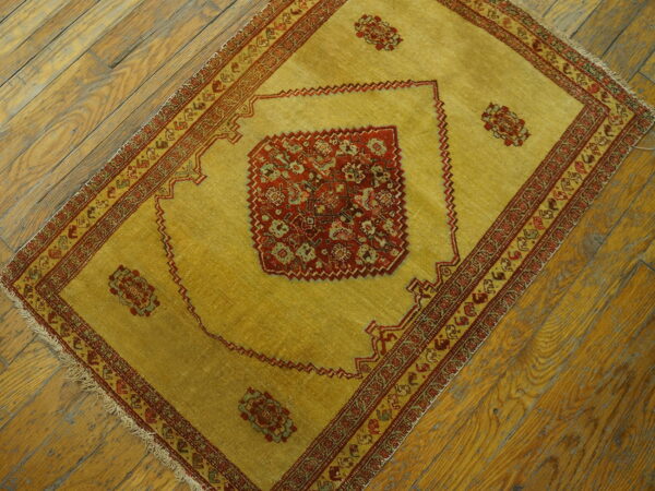 Low-pile yellow-gold rug with red traditional patterns and fringe rests on diagonal distressed wood planks.