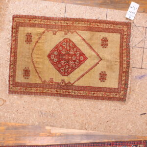 Golden-tan rug with a red central medallion and borders displayed on an OSB board over wood planks.