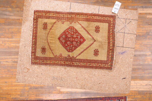 Golden-tan rug with a red central medallion and borders displayed on an osb board over wood planks.
