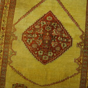 Low-pile rug with a gold field, red floral medallion, and geometric side borders on wooden floorboards.