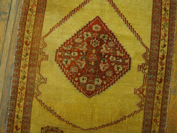 Low-pile rug with a gold field, red floral medallion, and geometric side borders on wooden floorboards.