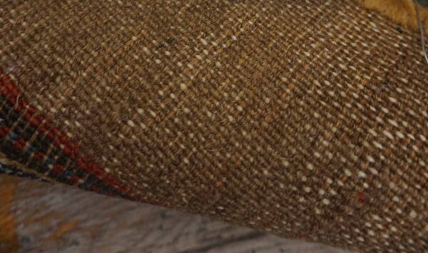 Close-up of a rug's brown woven backing with white flecks and visible red and blue pattern along the edge.