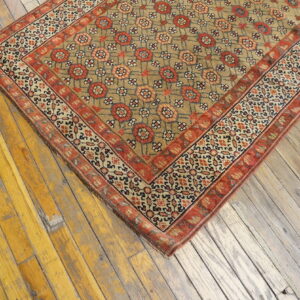 Corner of a low-pile tan and red geometric rug resting on distressed, wide-plank wood flooring.