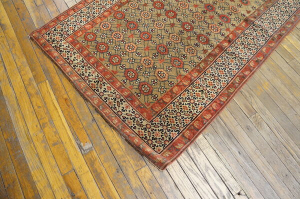 Corner of a low-pile tan and red geometric rug resting on distressed, wide-plank wood flooring.