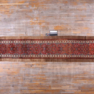 Rust-colored patterned runner rug with high brown edges lies on worn gray and orange wood flooring.