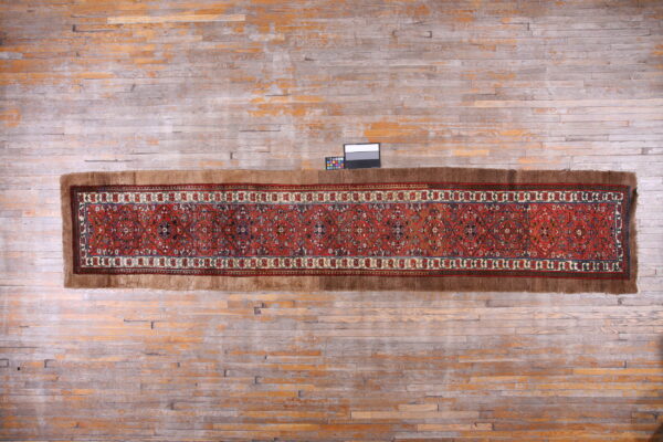Rust-colored patterned runner rug with high brown edges lies on worn gray and orange wood flooring.