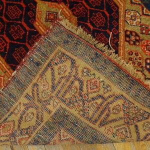 The corner of a geometric red, navy, and khaki rug is folded over, exposing its tight woven back on wood flooring.