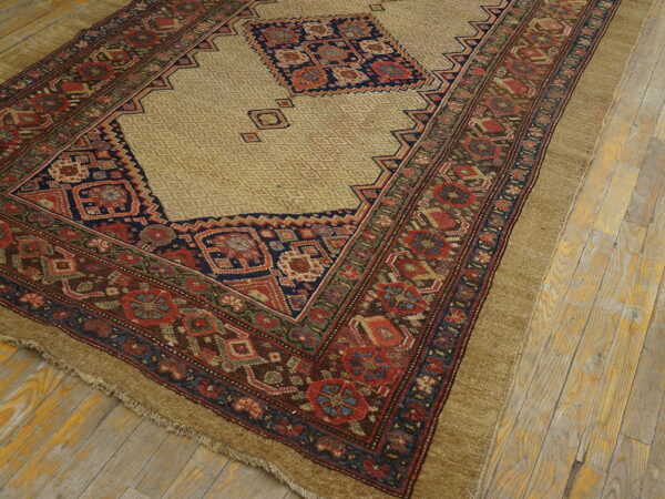 Geometric tan rug with red, navy, and brown complex borders lies on distressed light wooden floors.