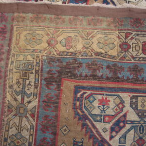 Geometric patterned rug corner in tan, blue, dark brown, and red resting on worn wood planks.