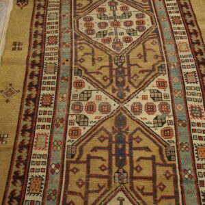Low-pile rug with intricate geometric patterns in gold, tan, red, and slate blue on distressed wood flooring.