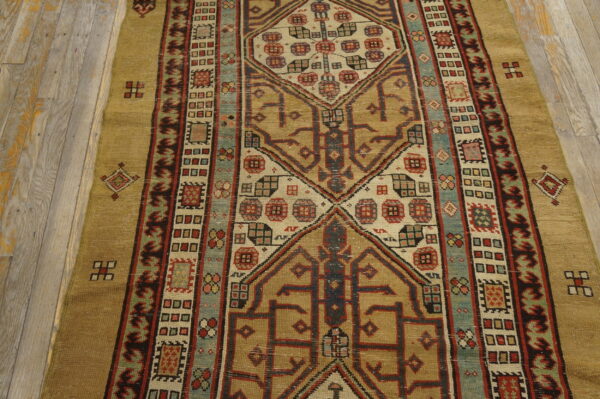 Low-pile rug with intricate geometric patterns in gold, tan, red, and slate blue on distressed wood flooring.