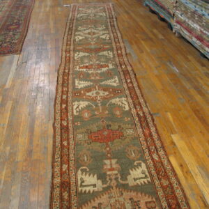 A sage green and rust geometric runner lies on worn hardwood floors near a stack of colorful rugs.