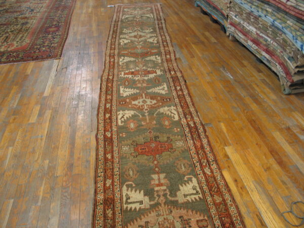 A sage green and rust geometric runner lies on worn hardwood floors near a stack of colorful rugs.