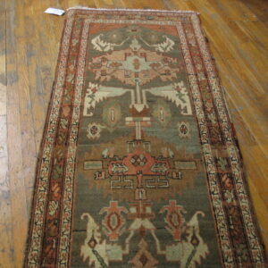 Geometric runner rug in muted olive green, rust, and tan displayed on worn hardwood flooring.