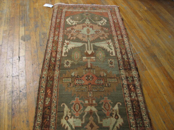Geometric runner rug in muted olive green, rust, and tan displayed on worn hardwood flooring.