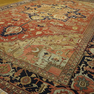 Ornate traditional rug with a rust field, deep navy border, and beige accents on wood floorboards.