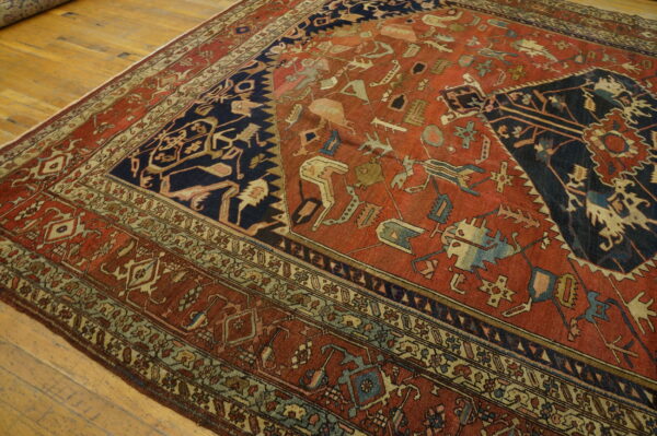 Geometric red and dark blue rug featuring stylized motifs and multiple borders on a light wood floor.