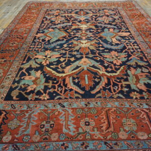 Low pile rug with navy blue, rust, and teal complex motifs sits on aged light wood flooring.
