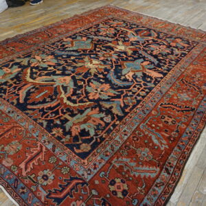 A large navy and red ornate rug is displayed on worn wood planks, next to white storage bags.