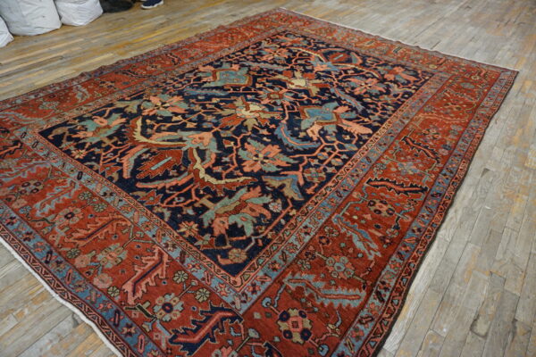 A large navy and red ornate rug is displayed on worn wood planks, next to white storage bags.