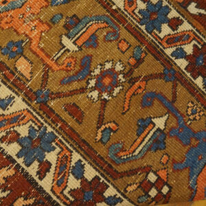 Close-up of a rug with a camel field, rust, blue, and orange geometric and floral motifs on wood flooring.