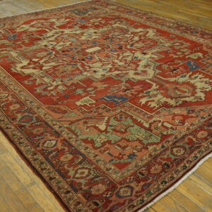 Ornate red rug with complex geometric patterns in cream, sage green, and blue on distressed wood flooring.