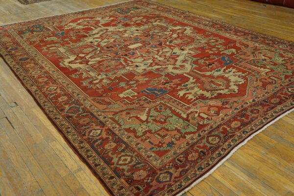 Ornate red rug with complex geometric patterns in cream, sage green, and blue on distressed wood flooring.
