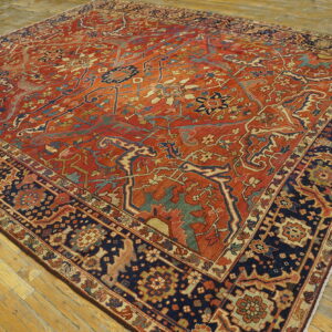 Ornate rug featuring a rust-red field, navy border, and complex floral motifs rests on light hardwood planks.