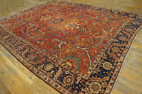 Ornate rug featuring a rust-red field, navy border, and complex floral motifs rests on light hardwood planks.