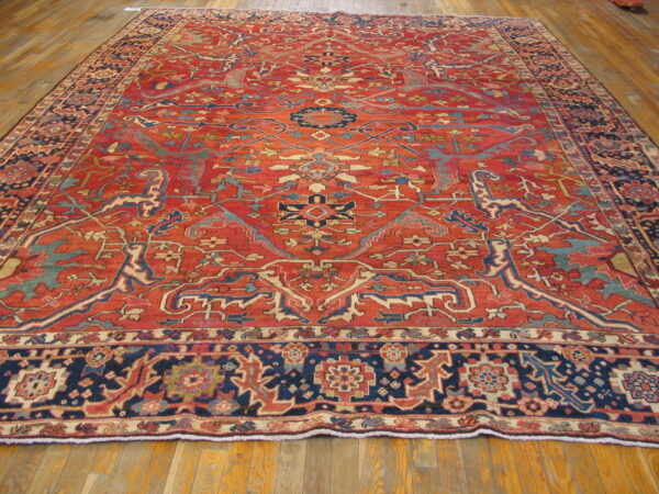 Large traditional rug with intricate red, navy, and teal patterns covering a light brown wood floor.