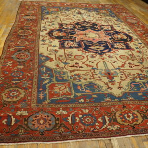 Traditional area rug featuring a navy and coral medallion on cream, bordered in red, sitting on wood floors.