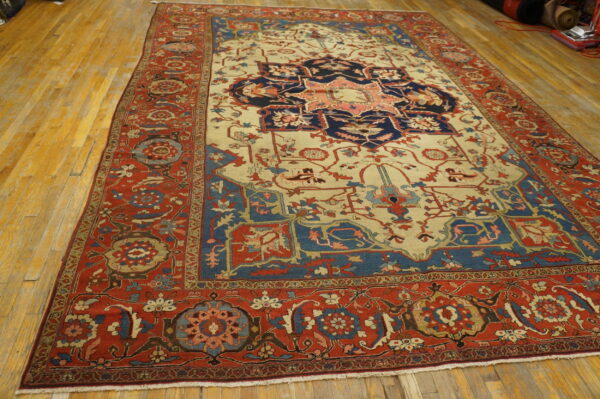 Traditional area rug featuring a navy and coral medallion on cream, bordered in red, sitting on wood floors.