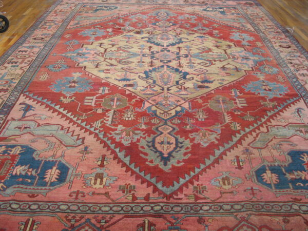 Oversize rug with a large beige geometric diamond framed by red, pink, and blue designs on wood floors.