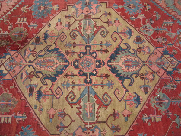 A traditional rug featuring a geometric medallion in khaki, blue, and muted pink on a reddish-maroon field.
