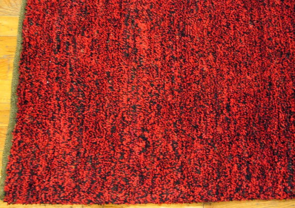 Red and black mottled, high-pile rug on light wood flooring, showing a green binding edge.