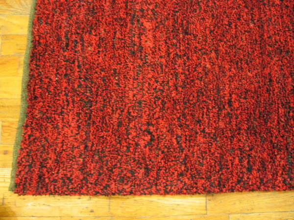Red and black thick pile rug with an olive green edge rests on light wood plank flooring.