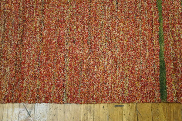 Textured rug with variegated red, orange, and yellow pile features a narrow vertical green stripe on wood.
