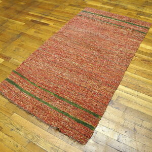Speckled red and orange textured rug with two green horizontal stripes rests on old plank hardwood.
