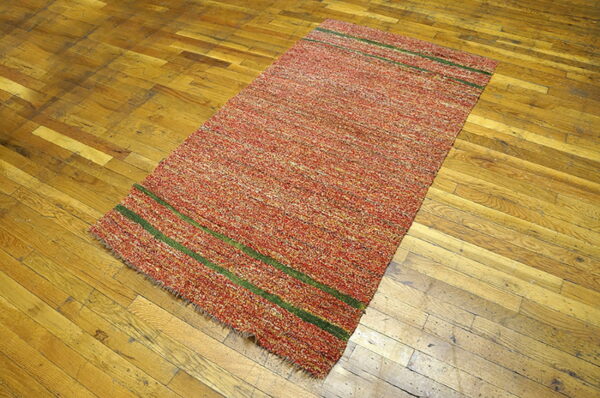 Speckled red and orange textured rug with two green horizontal stripes rests on old plank hardwood.
