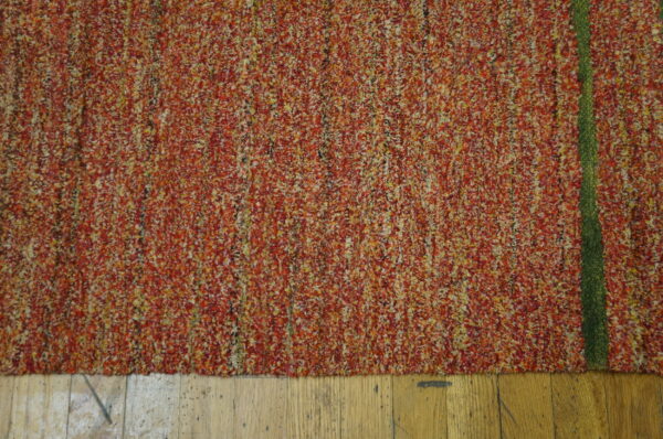 Textured, multi-tonal orange and red rug with a prominent green stripe, laid on wood flooring.