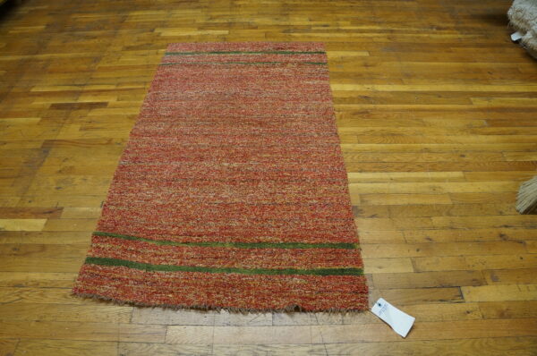 Textured red and orange mélange rug with two thin green stripes rests on distressed wood plank flooring.