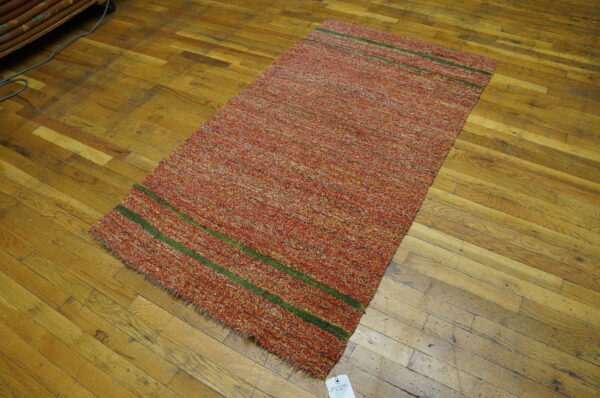 Textured red and yellow rug featuring two green stripes lies on worn, golden-toned wood floors.