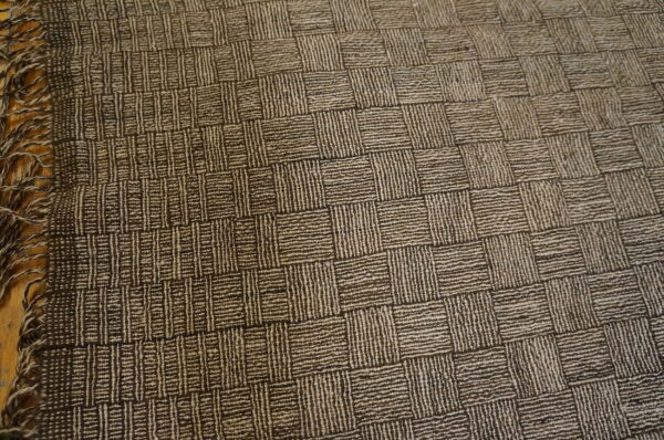 Textured dark brown and tan checkerboard rug with visible fringe lying on a wood surface.